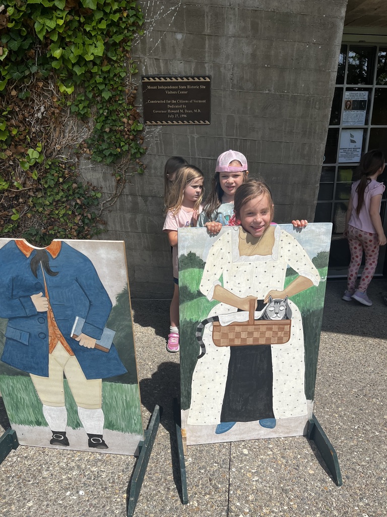 A young girl makes a funny, crinkled-face expression while posing behind a photo cutout board of a colonial woman holding a cat in a basket. Next to her, an empty cutout board of a man in a blue coat stands unused. In the background, two other children look on.  