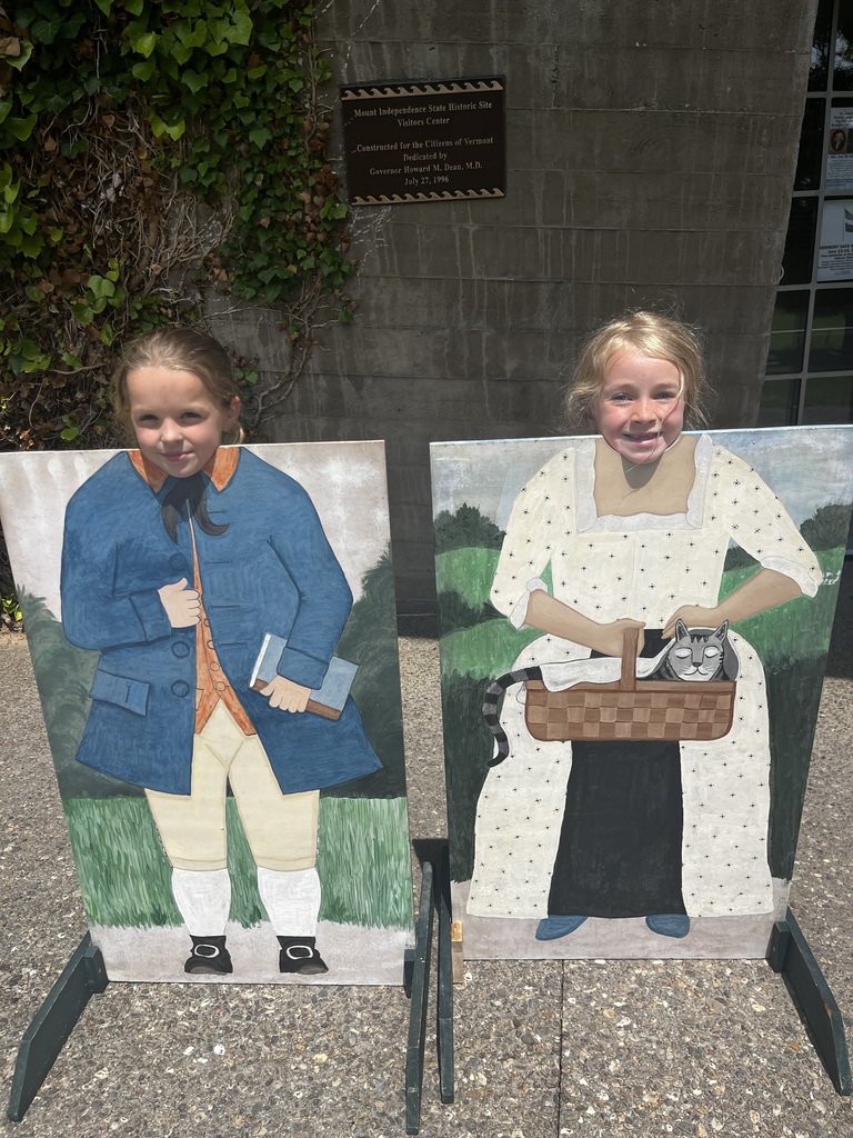 Two young girls smile warmly while posing behind historical photo cutout boards on a sunny day. The girl on the left stands behind the cutout of a colonial man holding a book, while the girl on the right stands behind the cutout of a colonial woman holding a cat in a basket. 