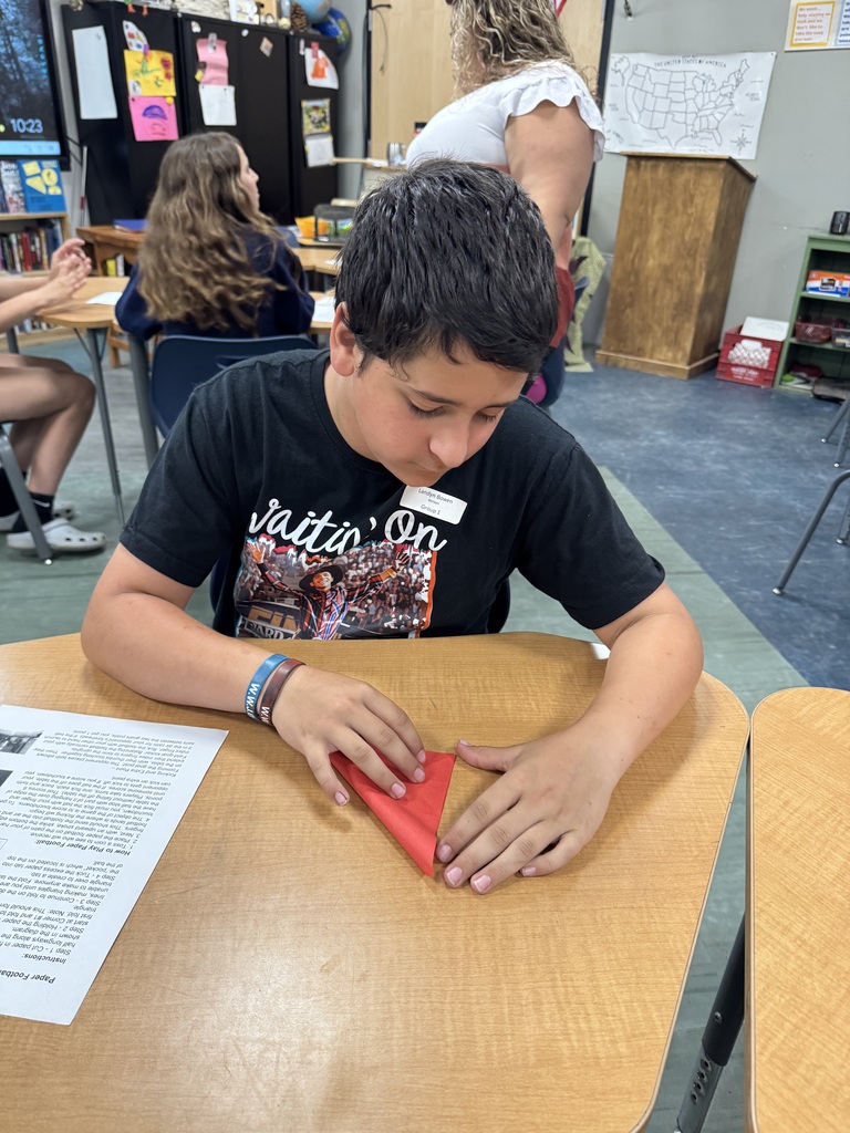 A close-up of a boy with short dark hair sitting at a school desk, looking down intently as he carefully folds a piece of red paper into a triangle shape. An instructional sheet for making a paper football is visible on the desk to his left. He is wearing a black graphic t-shirt and two wristbands.  IMG_5959.jpg: A girl with long brown hair and glasses sitting at a school desk, smiling as she looks down at a small piece of purple paper she is holding in both hands. An instructional sheet titled "Paper Football Game" sits on the desk in front of her. Other students and adults are visible in the background of the classroom.