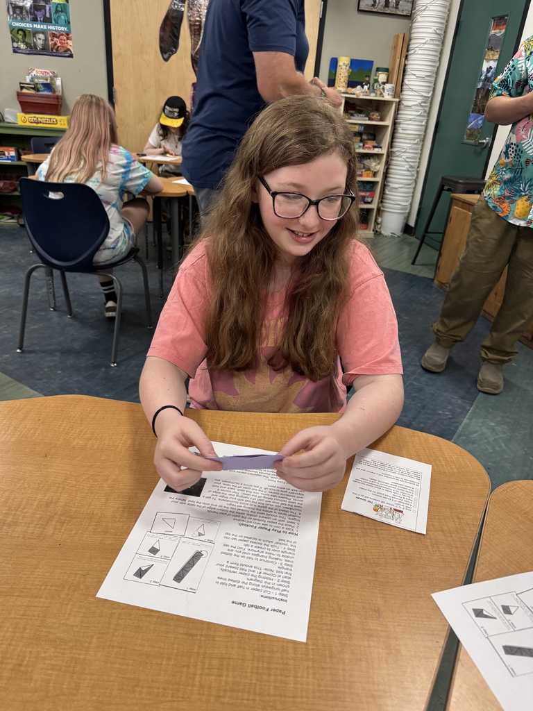  A girl with long brown hair and glasses sitting at a school desk, smiling as she looks down at a small piece of purple paper she is holding in both hands. An instructional sheet titled "Paper Football Game" sits on the desk in front of her. Other students and adults are visible in the background of the classroom.