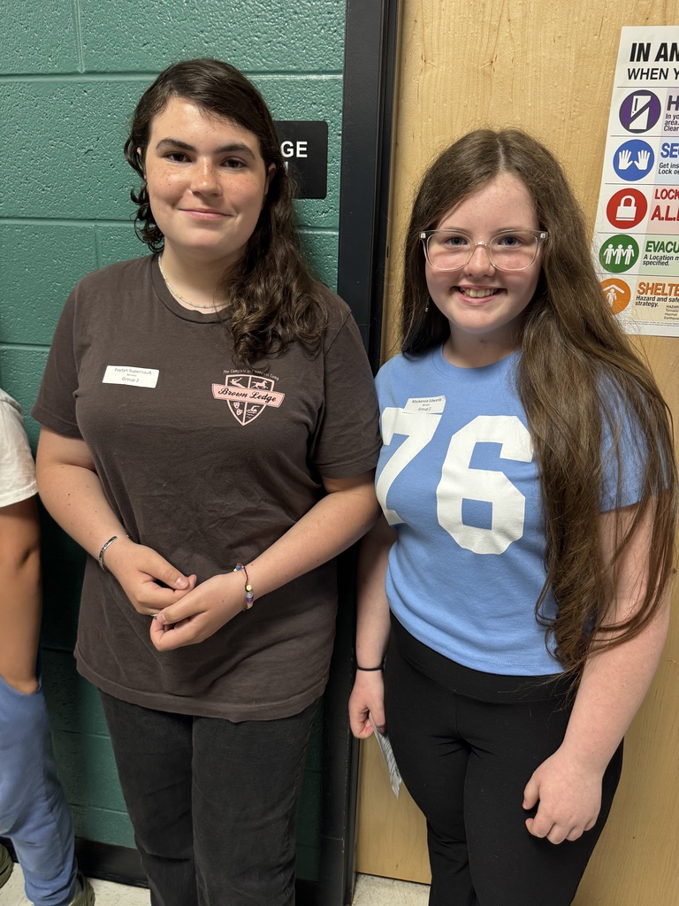 : Two young girls standing side-by-side indoors against a green cinderblock wall and a light wood door. The girl on the left has shoulder-length brown wavy hair and wears a dark brown t-shirt with a pink logo. The girl on the right has long brown hair, wears clear-rimmed glasses, and a light blue t-shirt with the number "76" printed on the front. Both are smiling at the camera and wearing small white name tags. 