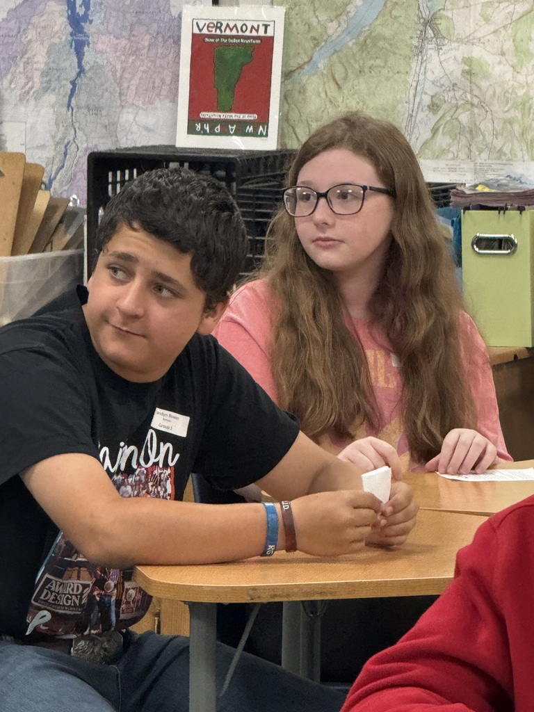 Two students are seated next to each other at a desk in a classroom. On the left, a boy in a black graphic t-shirt looks off to the side with a slight smirk, holding a small piece of white paper. Next to him, a girl with long wavy brown hair and glasses looks forward with a neutral expression, wearing a pink t-shirt. The background features a large map on the wall and a small sign that reads "VERMONT" above a green silhouette of the state.