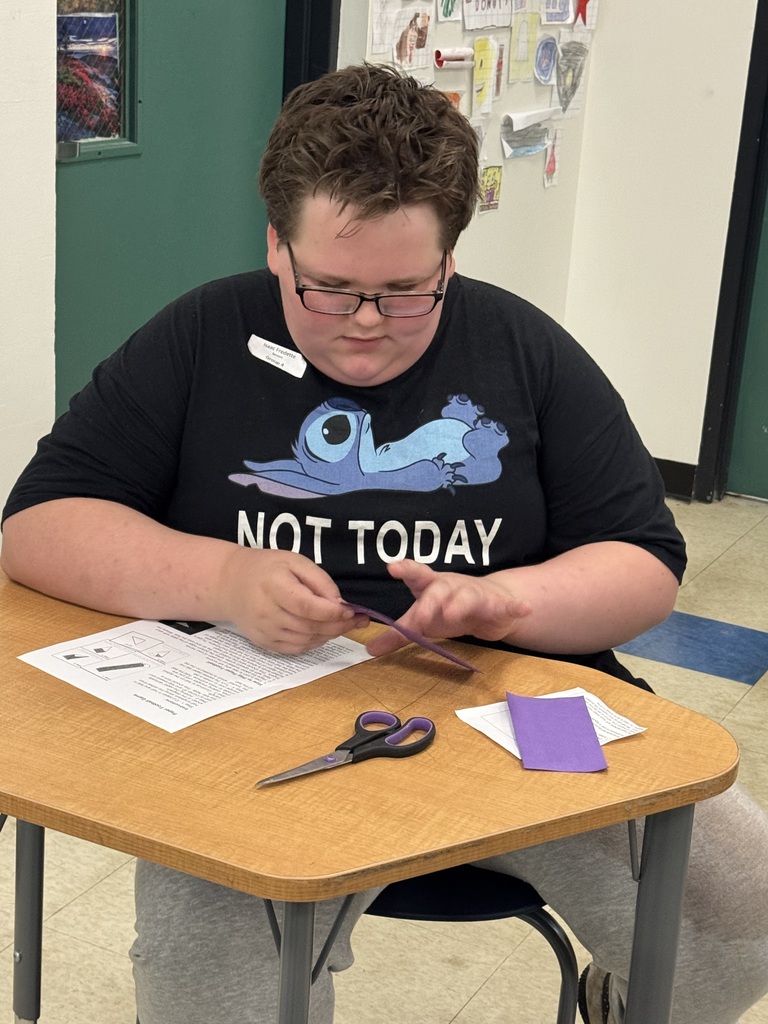  A boy with glasses and wavy brown hair is seated at a wooden classroom desk, looking down intently at a piece of purple paper he is folding. He is wearing a black t-shirt featuring the cartoon character Stitch and the text "NOT TODAY." On his desk, there is an instructional sheet, a pair of black and purple scissors, and another small piece of purple paper. In the background, a green door frame and student artwork displayed on the wall are visible.  