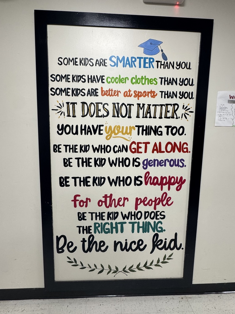 A vertical, framed motivational poster hangs on a beige classroom wall. The text uses a variety of colorful fonts and hand-lettered styles. It reads: "SOME KIDS ARE SMARTER THAN YOU. SOME KIDS HAVE cooler clothes THAN YOU. SOME KIDS ARE better at sports THAN YOU. IT DOES NOT MATTER. YOU HAVE your THING TOO. BE THE KID WHO CAN GET ALONG. BE THE KID WHO IS generous. BE THE KID WHO IS happy for other people. BE THE KID WHO DOES THE RIGHT THING. Be the nice kid." A small illustration of a blue graduation cap sits at the top, and a simple green leafy vine illustration decorates the bottom.   