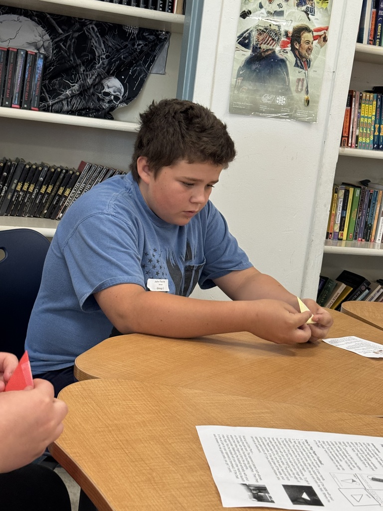 A young boy with short brown hair sits at a light-colored wooden school desk, leaning forward with his elbows resting on the surface. He is wearing a blue t-shirt and a small white name tag on his chest. His attention is focused downward on a yellow piece of paper he is holding and folding with both hands. In front of him on the desk is an instructional worksheet titled "Paper Football Game." The background shows a classroom setting with bookshelves filled with novels and a hockey-themed poster on the wall. Another student's hands holding red paper are partially visible in the bottom-left corner. 