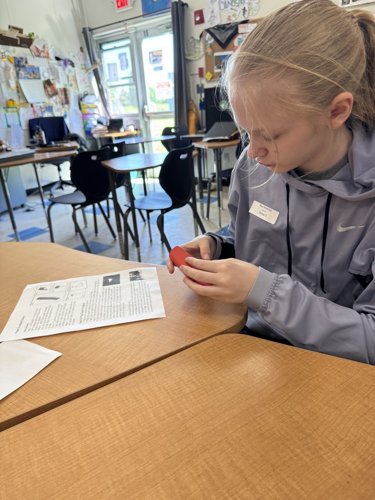 The same young girl with blonde hair in a ponytail is shown from a similar angle, but she is now looking down, focused on carefully folding the red paper in her hands. The instructional sheet for the "Paper Football Game" sits flat on the wooden desk in front of her. The classroom background remains the same, showing desks and a sunlit door.