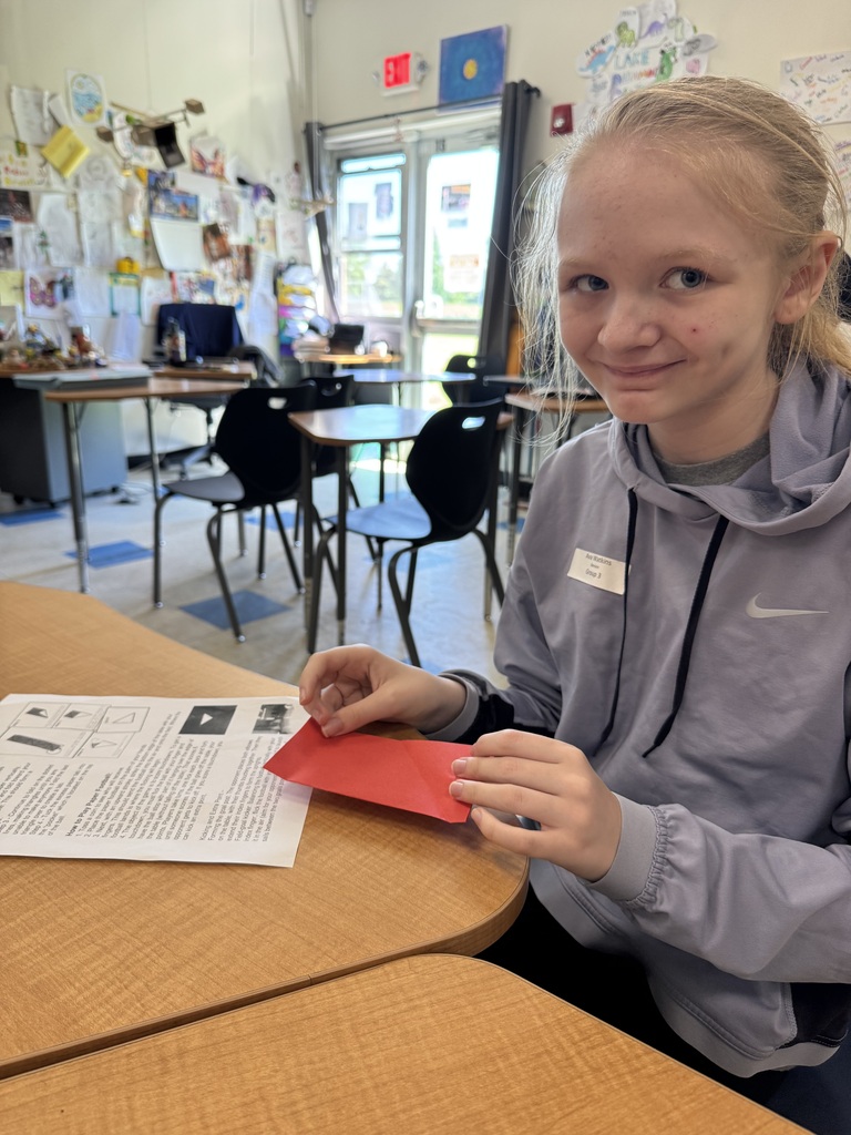 A young girl with blonde hair tied back in a ponytail sits at a wooden desk, smiling directly at the camera. She is wearing a grey Nike hooded sweatshirt with a name tag. She holds a folded piece of red paper over an instructional sheet on her desk. The background shows a bright classroom with empty desks, a glass door leading outside, and walls decorated with various student projects.