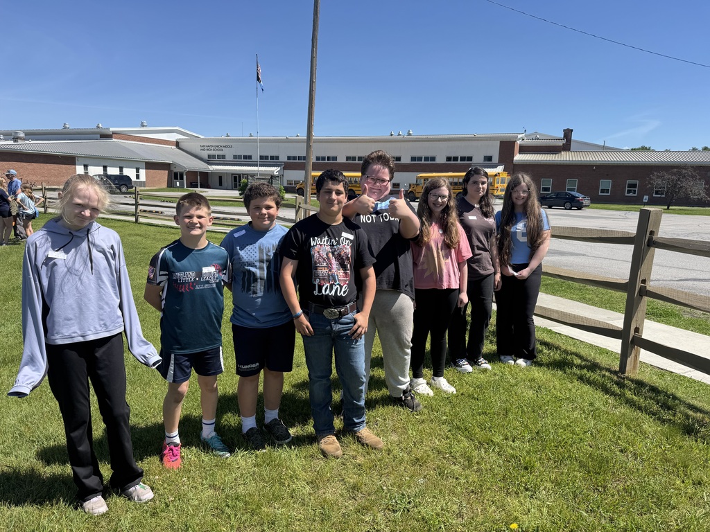 A group photo of seven diverse children and teenagers lined up on a vibrant green lawn on a clear, sunny day. They are dressed in casual clothes, t-shirts, and sneakers, with a few of them smiling or giving a thumbs-up. In the background is a long, brick and white school building under a bright blue sky, with an American flag flying on a tall pole. Yellow school buses and a couple of cars are parked in the driveway behind them, and a rustic wooden fence frames the right side of the lawn.