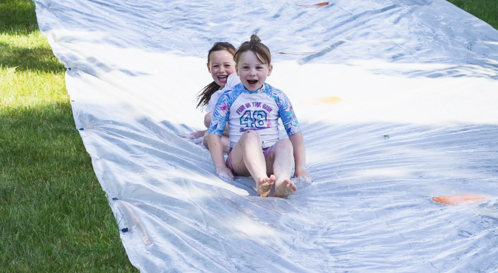 Two elementary students laugh as they slide together down a large slip-and-slide during an outdoor end-of-year celebration.