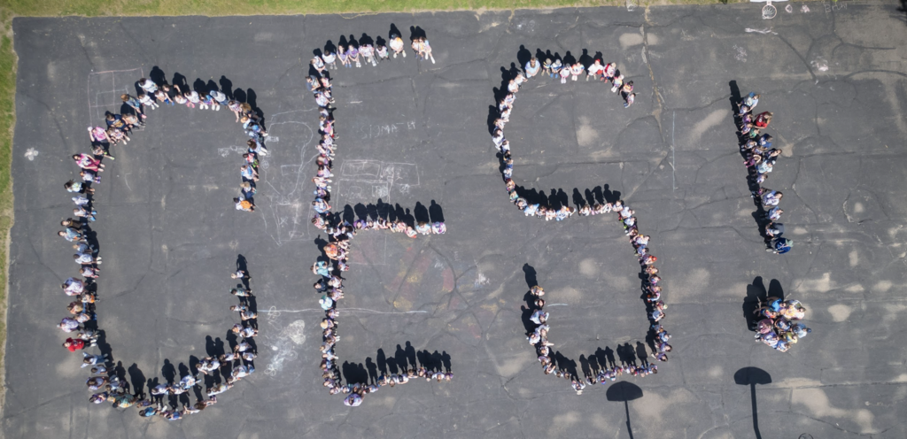 A drone photograph shows Castleton Elementary School students and staff standing on the blacktop arranged to spell the letters "CES" with an exclamation point, demonstrating teamwork and school pride.