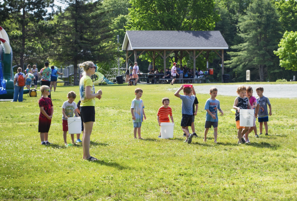 Students participate in an outdoor game on a grassy field while staff members supervise. A pavilion, playground equipment, and other celebration activities are visible in the background.