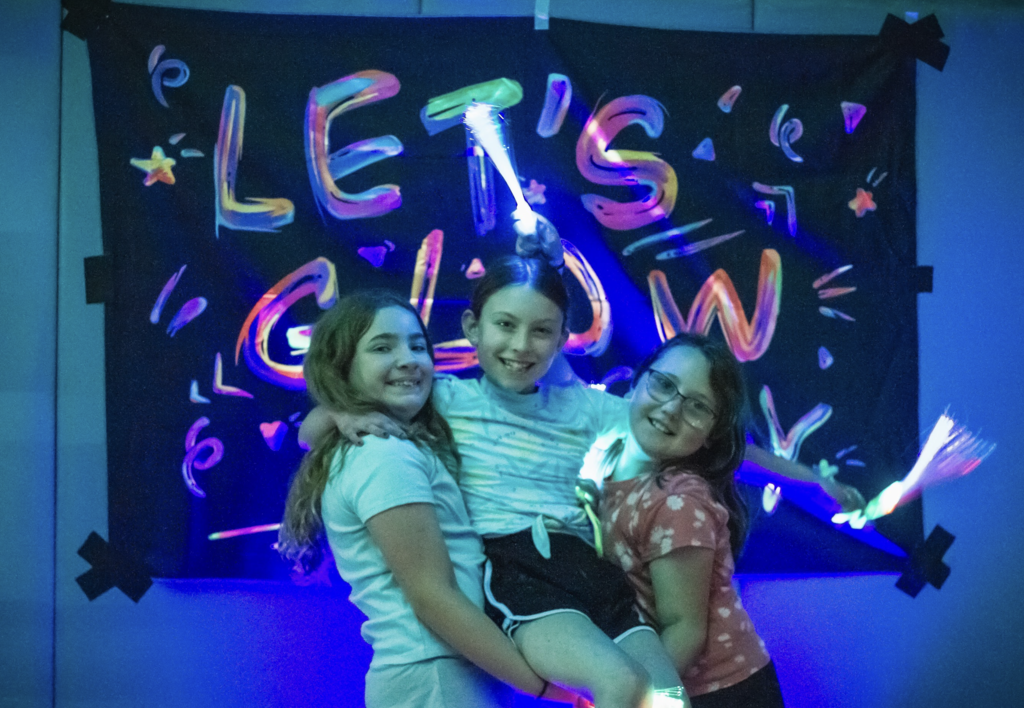 Three students pose together in front of a glowing blacklight backdrop that reads "Let's Glow," holding light-up fiber optic wands and smiling for the camera.