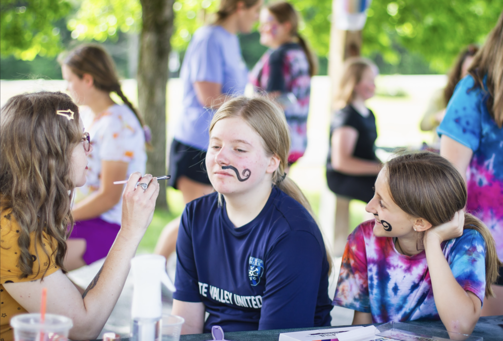 A student receives face paint while sitting with friends at a picnic table. One student has a painted mustache design on their face.