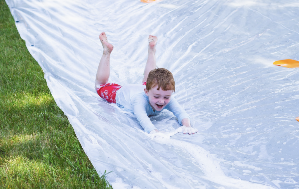 A young student wearing a white rash guard and red swim shorts smiles while sliding down a soapy slip-and-slide during Castleton Elementary School's Big Celebration.