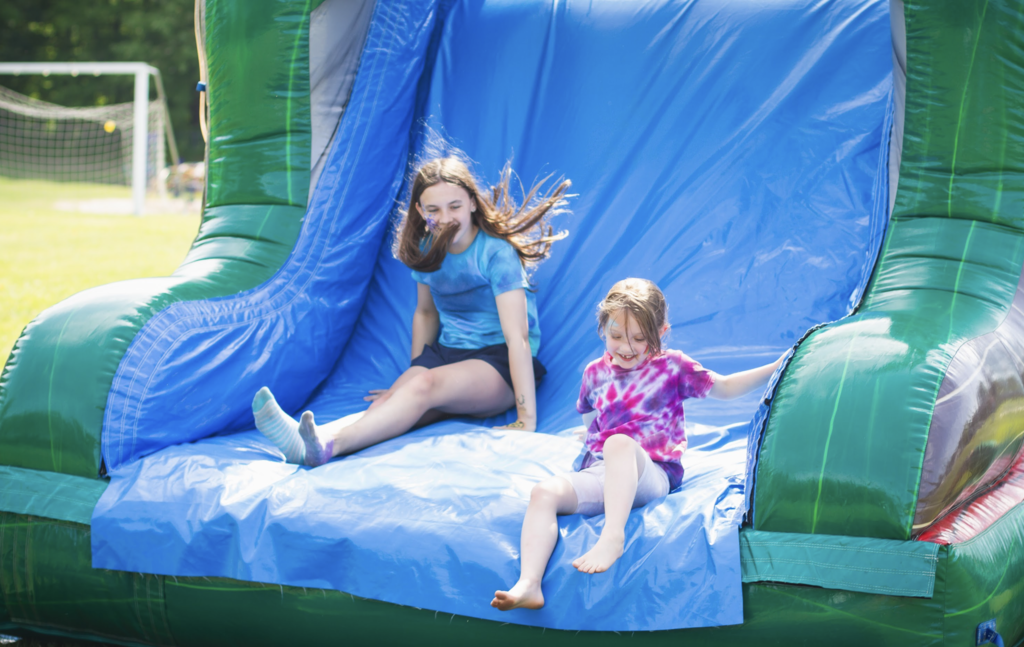 Two students smile as they slide down a large blue inflatable slide during the outdoor celebration.