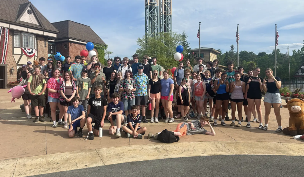 A large group of middle and high school students pose together outside an amusement park entrance on a sunny day. The group stands in front of a building decorated with patriotic bunting and several red, white, and blue balloon arrangements. Some students hold stuffed animals and prizes, while others wear colorful shirts and summer attire. A tall amusement park ride tower rises in the background, and several students smile and gesture toward the camera during the group photo.