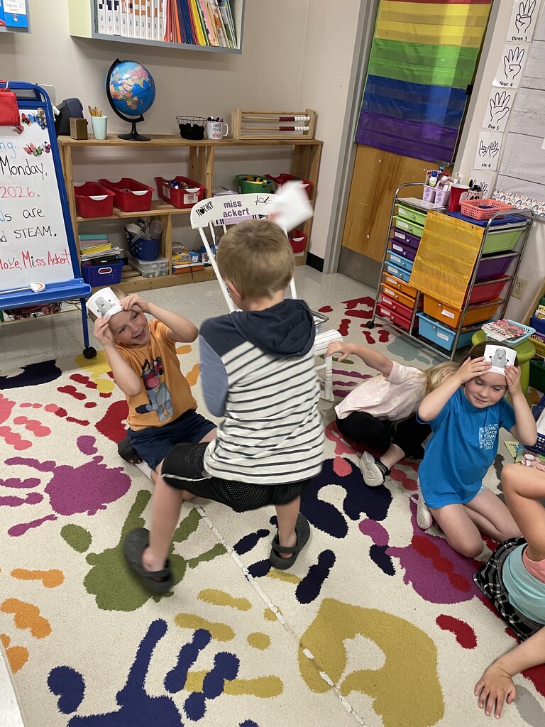 A group of six young children are sitting in a circle on a vibrant, multi-colored handprint-patterned rug in a classroom. Each child is holding a square white card featuring a cartoon mole illustration against their forehead. A classroom whiteboard in the background displays a message written in marker: "Today is Monday, June 8, 2026. Our specials are health and STEAM. Love, Miss Ackert." A white rocking chair with "miss ackert" written on the backrest sits in the background near colorful storage organizers. .