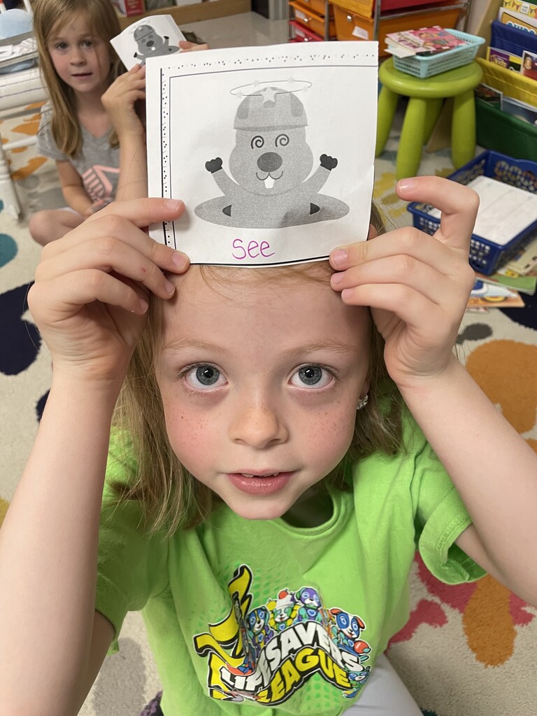 A close-up shot of a young girl with blue eyes and light brown hair with freckles, wearing a bright green t-shirt. She is sitting on a colorful classroom rug and holding a square white card against her forehead with both hands. The card features a black-and-white cartoon illustration of a mole popping out of a hole with dizzy swirls in its eyes and stars spinning over its construction hat. The word "See" is handwritten in pink marker at the bottom of the card. In the background, another young girl is partially visible, holding up a similar card.