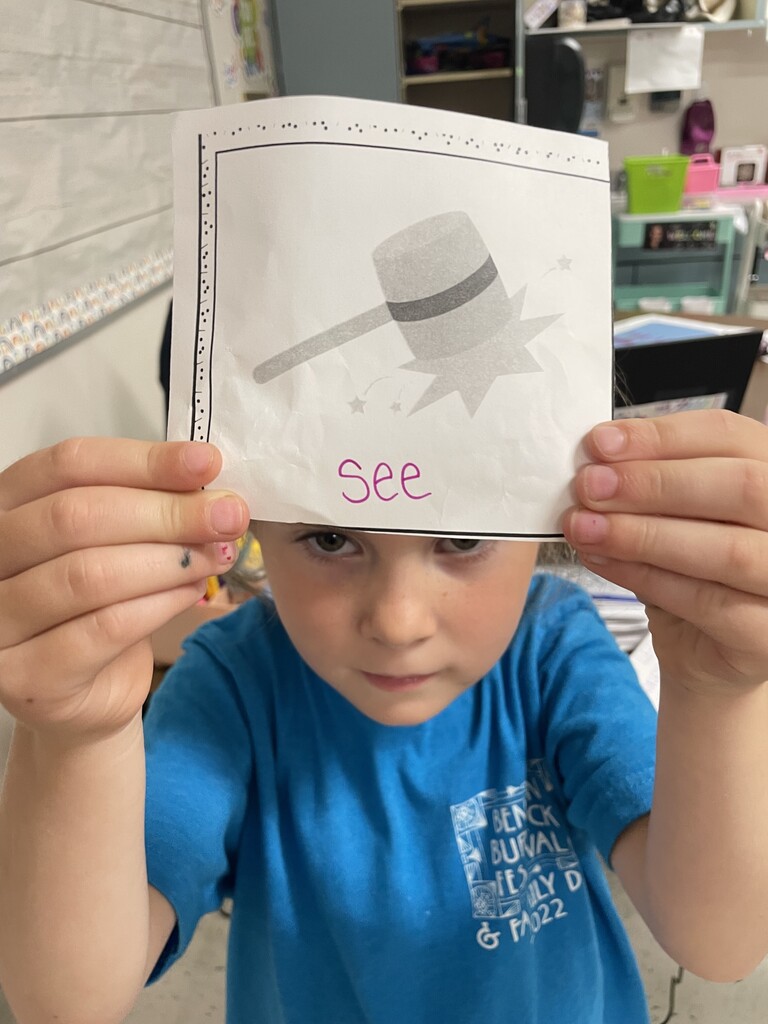 A close-up shot focusing on a young girl wearing a bright blue t-shirt, who is holding a white card directly over her forehead and eyes. The card shows a gray cartoon illustration of a mallet striking a surface with impact sparks and stars. Below the image, the word "See" is written in pink marker. The background shows a blurred classroom setting with storage bins and a colorful rainbow-patterned wall border.