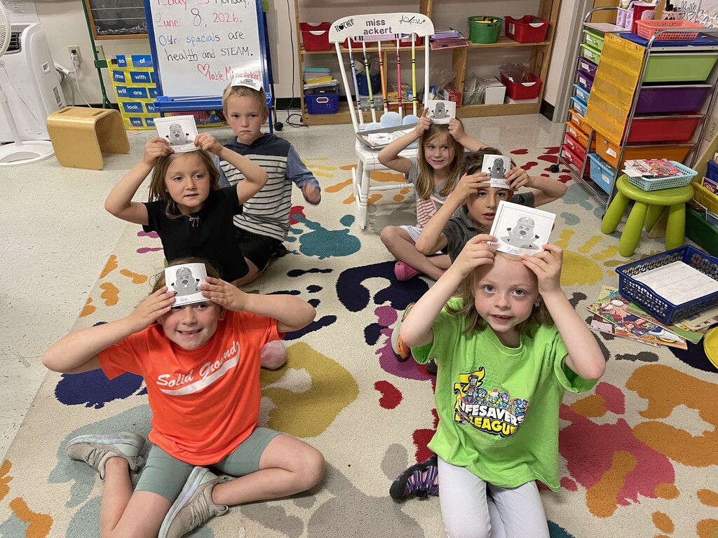 A group of six young children are sitting in a circle on a vibrant, multi-colored handprint-patterned rug in a classroom. Each child is holding a square white card featuring a cartoon mole illustration against their forehead. A classroom whiteboard in the background displays a message written in marker: "Today is Monday, June 8, 2026. Our specials are health and STEAM. Love, Miss Ackert." A white rocking chair with "miss ackert" written on the backrest sits in the background near colorful storage organizers.