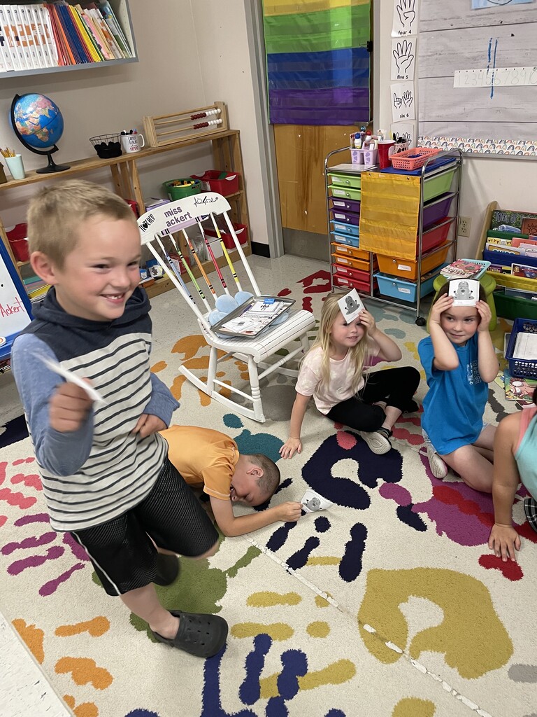 A classroom scene showing a group of young children playing on a colorful handprint rug. In the foreground, a young boy in a grey-and-white striped hoodie and black shorts smiles broadly as he steps forward. Behind him, another boy in an orange t-shirt lies on his stomach on the rug, looking at a cartoon card. To the right, two young girls are seated on the floor; one is holding a cartoon card to her forehead, and the other looks on. The classroom background includes a white rocking chair, shelves, a globe, and rainbow decor. IMG_6694.jpg An action shot in a classroom setting where several young children are playing on a colorful handprint rug. In the center, a boy in a striped hoodie is captured mid-movement, facing away from the camera. To his left, a boy in an orange shirt sits on the floor, laughing while holding a white card to his forehead. To the right, a girl in a blue shirt also holds a card to her forehead, while another girl sits nearby. The background features a white rocking chair, a globe on a wooden shelf, and a whiteboard with handwritten classroom schedule details.