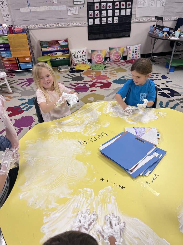 A high-angle shot looking down at a large yellow table in a classroom where children are playing with white shaving cream. Two young girls are visible; one with blonde hair smiles broadly toward the camera, lifting her foam-covered hands, while a girl in a bright blue shirt next to her rubs the foam onto the table surface. Handprints and upside-down names like "William" and "Demila" are written on the table near a stack of blue folders. The background shows a colorful classroom rug and cubbies.