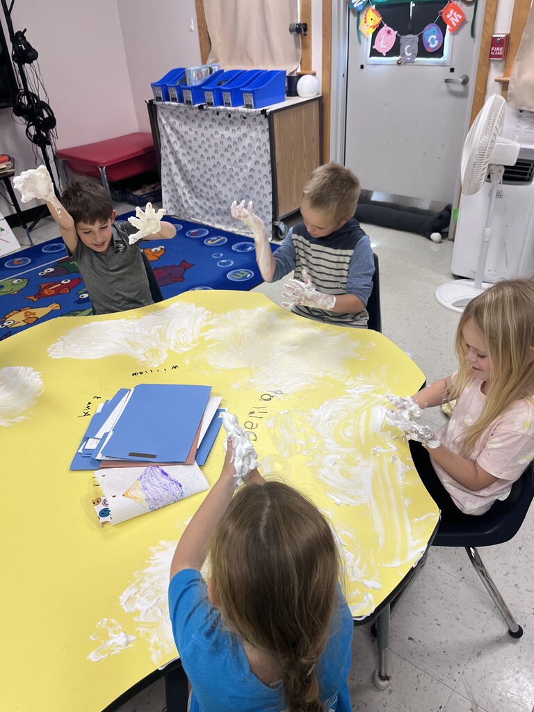 An overhead shot of four young children sitting around a large yellow, flower-shaped table in a classroom, enthusiastically playing with white shaving cream spread across the surface. Two boys on the far side hold their messy, foam-covered hands high in the air with big smiles. A girl with blonde hair on the right rubs the foam on the table, while another girl in a blue shirt in the foreground lifts her foam-covered hands. Stacked folders and a colorful drawing sit in the center of the table.