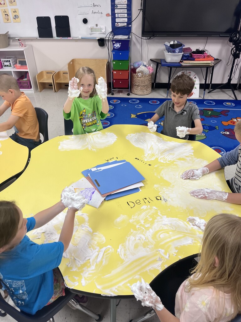 An overhead shot of four young children sitting around a large yellow, flower-shaped table in a classroom, enthusiastically playing with white shaving cream spread across the surface. Two boys on the far side hold their messy, foam-covered hands high in the air with big smiles. A girl with blonde hair on the right rubs the foam on the table, while another girl in a blue shirt in the foreground lifts her foam-covered hands. Stacked folders and a colorful drawing sit in the center of the table.