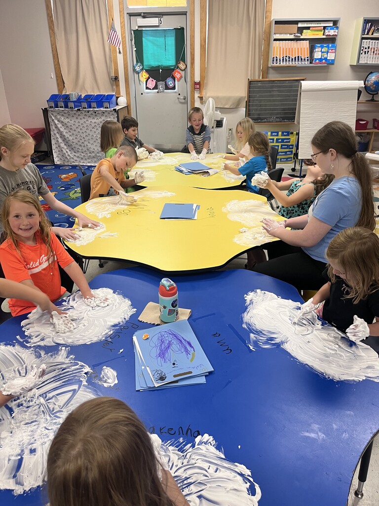 A wide shot of a classroom showing two large group tables pushed near each other—one bright yellow and one deep blue—with about ten young children and older students sitting around them. Everyone is actively engaged in sensory play, spreading white shaving cream all over the table surfaces. Several children laugh and hold up their messy hands, while others focus on smoothing the foam out in front of them. The classroom background features storage shelves, a whiteboard, and a window.