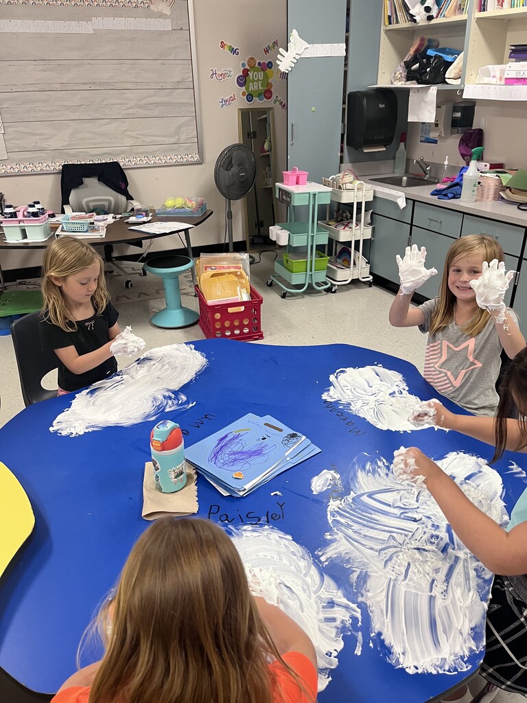 A high-angle shot of a classroom setting where children are sitting at a large, blue clover-shaped table covered in swirls of white shaving cream. A young girl with long blonde hair on the right smiles proudly at the camera, holding up both hands coated thick with foam. On the left, another girl concentrates on rubbing the shaving cream. Names like "Paisley" and "Makenna" are written on the table near a blue folder and a water bottle resting on a paper towel.