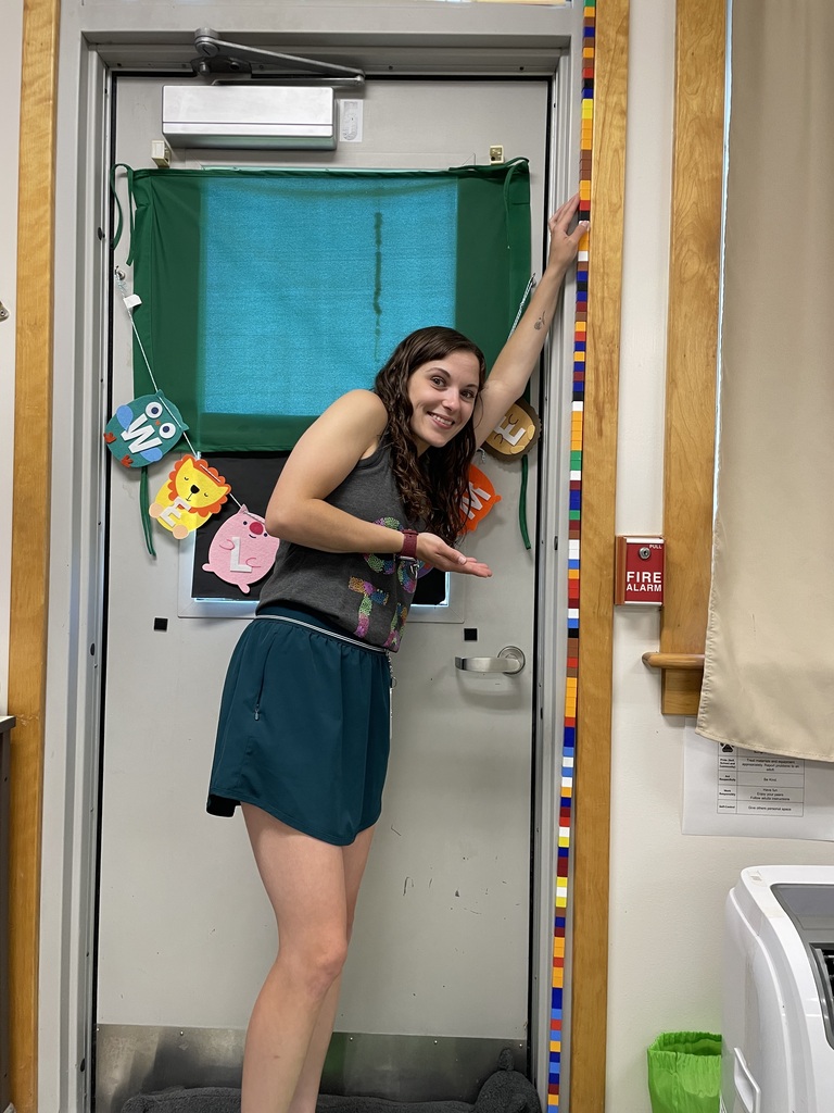A smiling woman with long brown hair stands in a classroom doorway, gesturing toward a tall vertical column of interlocking plastic blocks. She is wearing a grey tank top and dark green athletic shorts, and her left arm is raised to touch the top section of the block tower. The colorful tower of blocks is attached to the wooden door frame and stretches from the floor to the top of the door. The door behind her has a green privacy curtain and a colorful banner spelling out part of a word. A red fire alarm pull station is visible on the wall to the right.