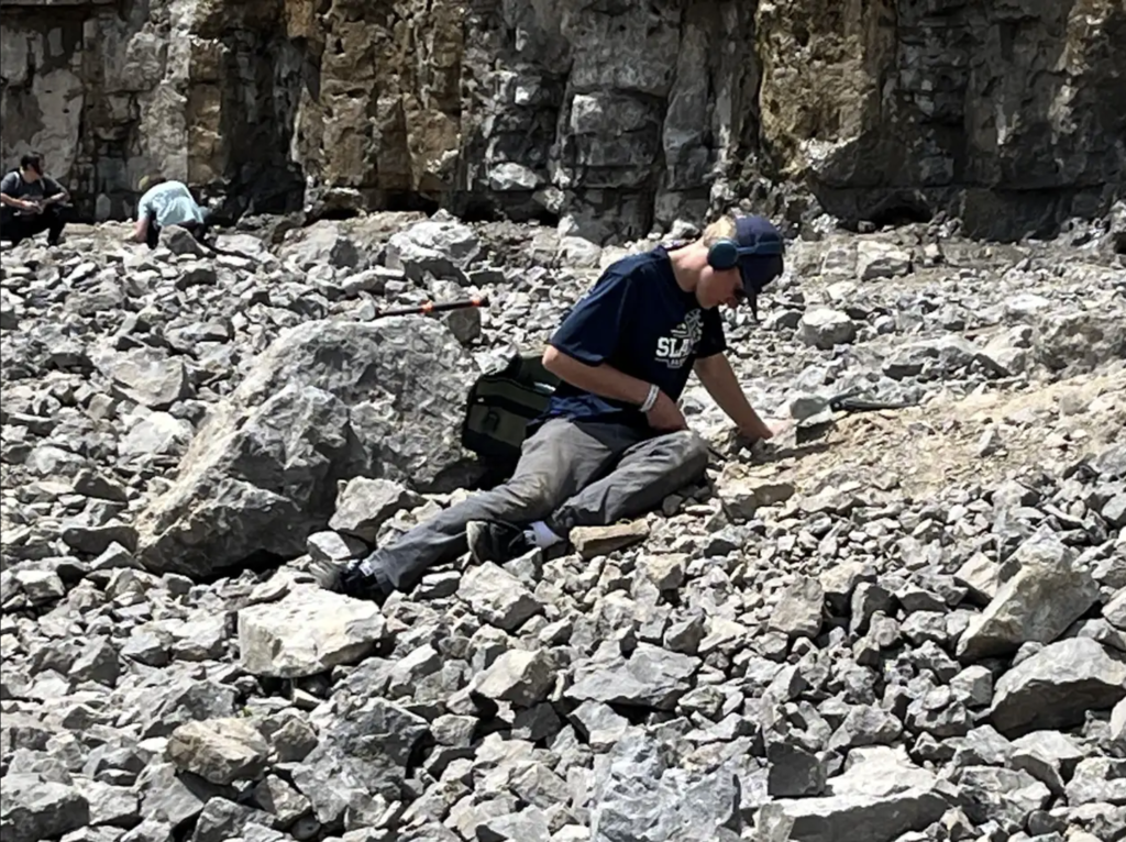 A student wearing safety glasses and earmuffs sits among rocks in the quarry, using a hand tool to carefully search through stone fragments for crystals.