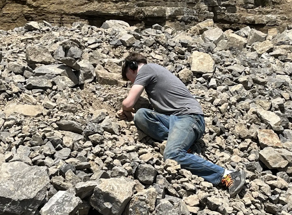 A student kneels on the rocky ground while chiseling into a section of stone, focused on uncovering potential mineral specimens.