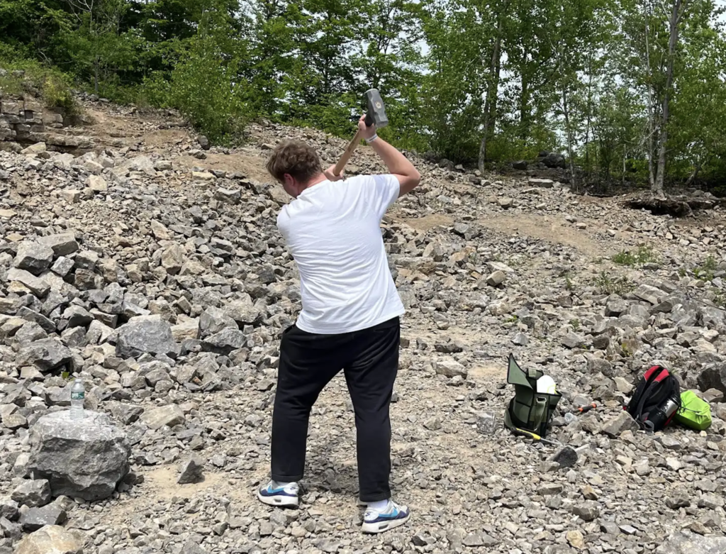 A student raises a sledgehammer overhead while standing among piles of stone in the quarry, ready to crack open a rock.