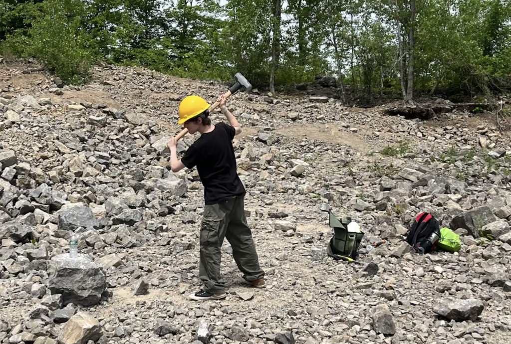 A student wearing a yellow hard hat stands in the quarry holding a large sledgehammer over one shoulder, preparing to break open rocks.