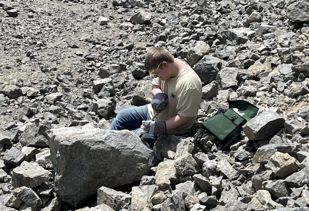 A student wearing safety glasses and work gloves uses a hammer and chisel beside a large boulder while searching for Herkimer Diamonds in the quarry.