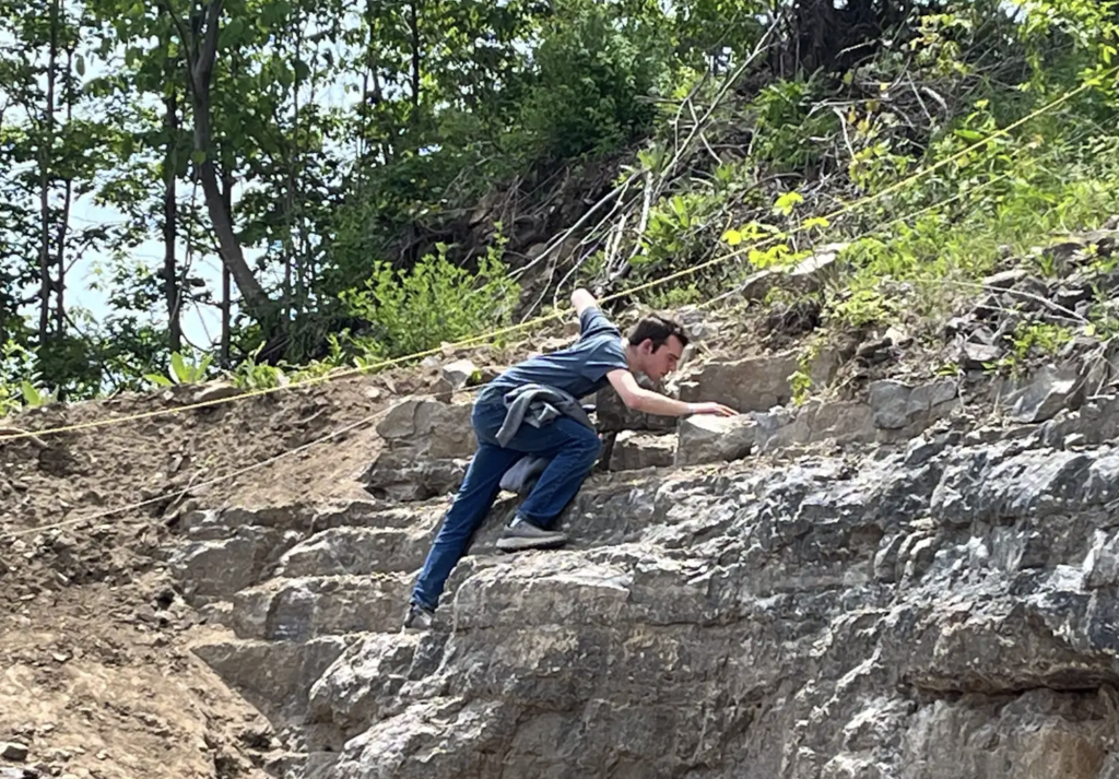 A student climbs across a rocky ledge and cliff face inside the quarry, exploring higher areas in search of mineral deposits.