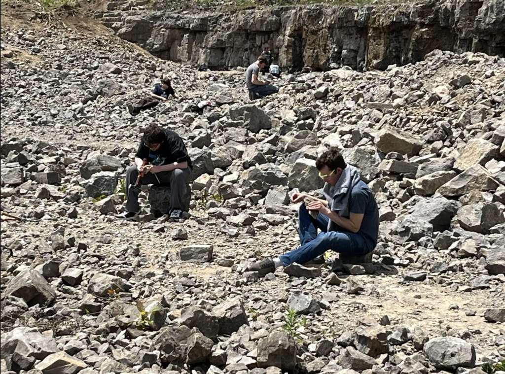 Several Honors Geology students sit and kneel among piles of broken rock in a large quarry while carefully searching for Herkimer Diamonds. A rocky cliff face rises behind them.
