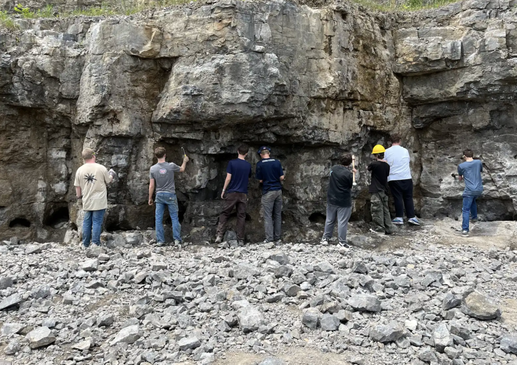 The Honors Geology class stands side by side along a rock wall in the quarry, using hammers and tools to search for Herkimer Diamonds in the exposed stone.