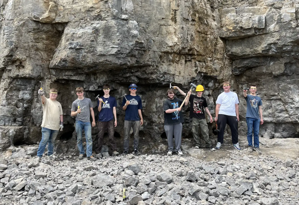 The Honors Geology class poses for a group photo in front of a large quarry rock face. Several students hold up hammers, chisels, and rock samples they collected during the trip.