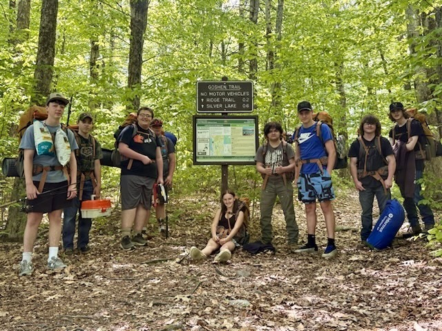 Outdoor Pursuits students pose with backpacks and camping equipment at the Goshen Trail trailhead sign surrounded by a green forest.