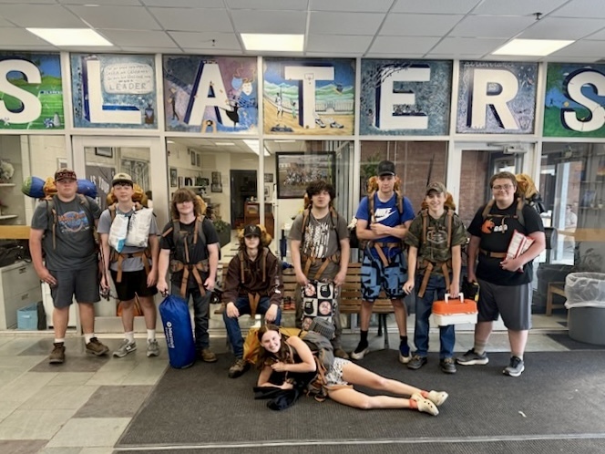 A group of Outdoor Pursuits students stands inside a school entrance with large backpacks, camping gear, sleeping pads, and supplies as they prepare to leave for their overnight trip to Silver Lake.