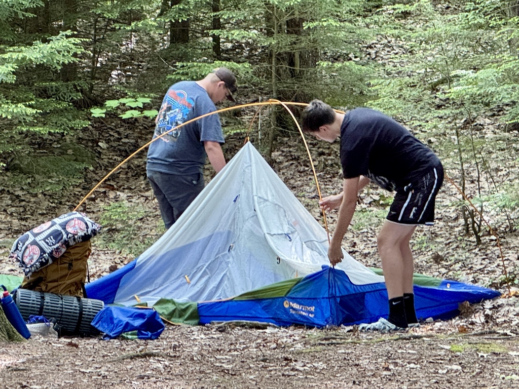 Two students work together to set up a backpacking tent in a wooded campsite surrounded by sleeping pads, backpacks, and outdoor equipment.