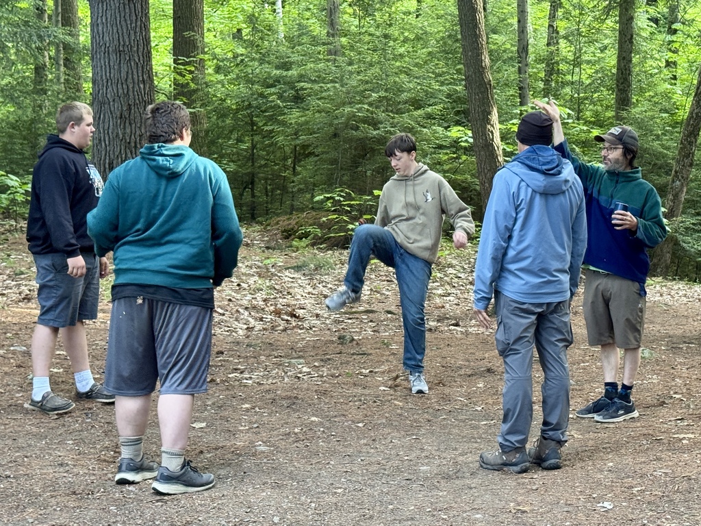 Students and an instructor participate in an Outdoor Pursuits Olympics activity in a forest clearing, standing in a circle while one student performs a challenge.