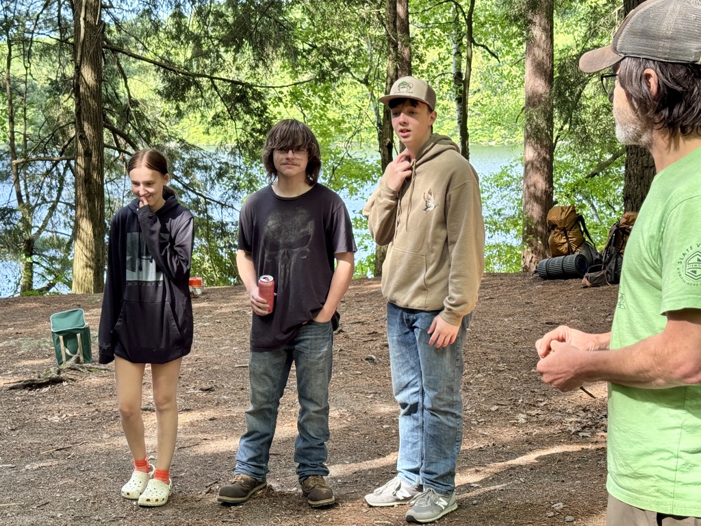 Three students stand in a forested campsite near Silver Lake listening during an Outdoor Pursuits activity while an instructor speaks nearby.