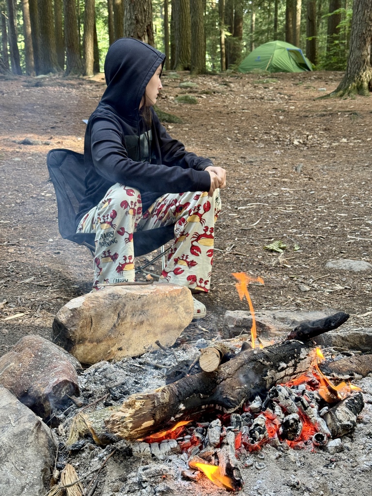 A student wearing a hoodie and pajama pants sits beside a campfire in the forest, warming up near glowing embers while a tent stands in the background.