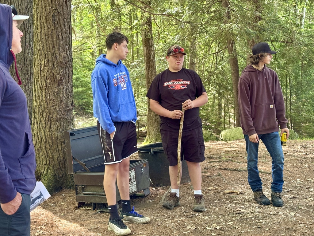 Students stand in a wooded campsite discussing a challenge during the Outdoor Pursuits Olympics, surrounded by tall trees and camping equipment.