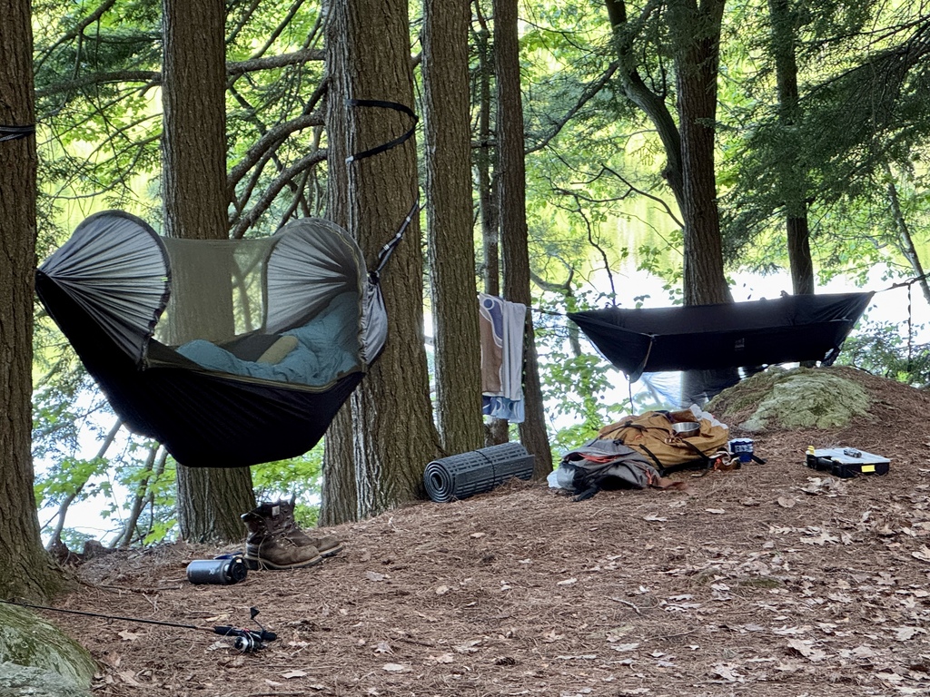 Two camping hammocks are suspended between tall trees at a wooded lakeside campsite. Backpacks, boots, and camping gear are arranged nearby.