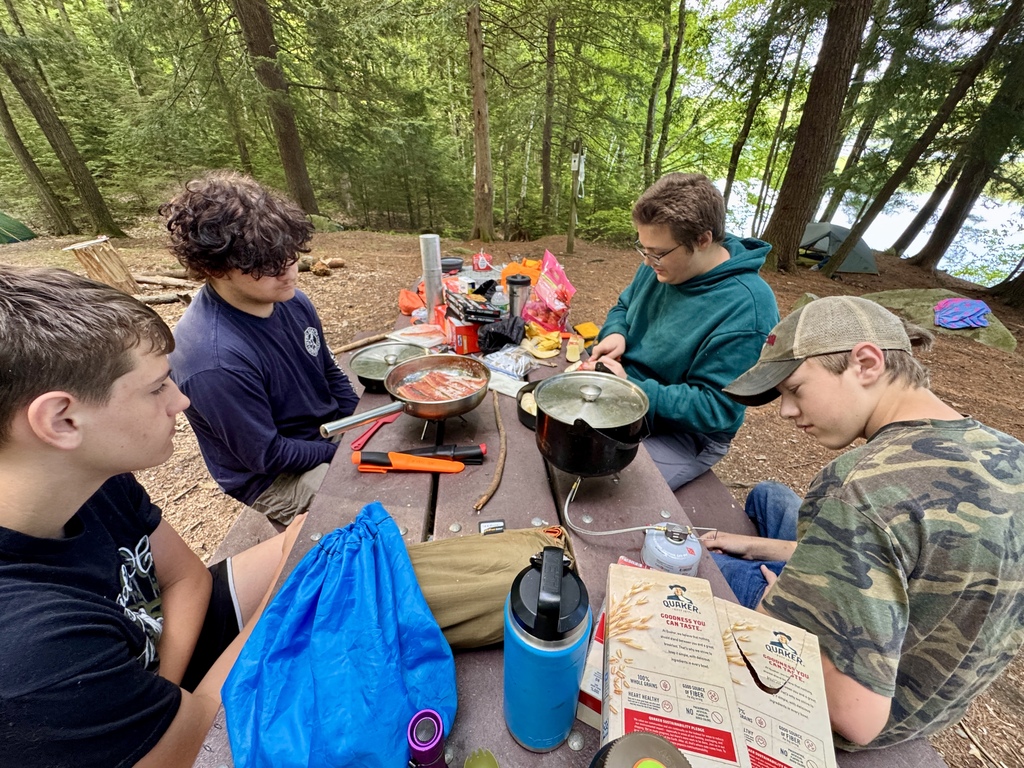 Students gather around a picnic table at their campsite preparing a meal using camp stoves and cookware, with food supplies and camping gear spread across the table.