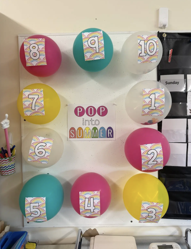 A classroom bulletin board displays colorful balloons numbered 1 through 10 surrounding a sign that reads “Pop Into Summer,” representing a countdown to the end of the school year with daily surprise activities hidden inside each balloon.